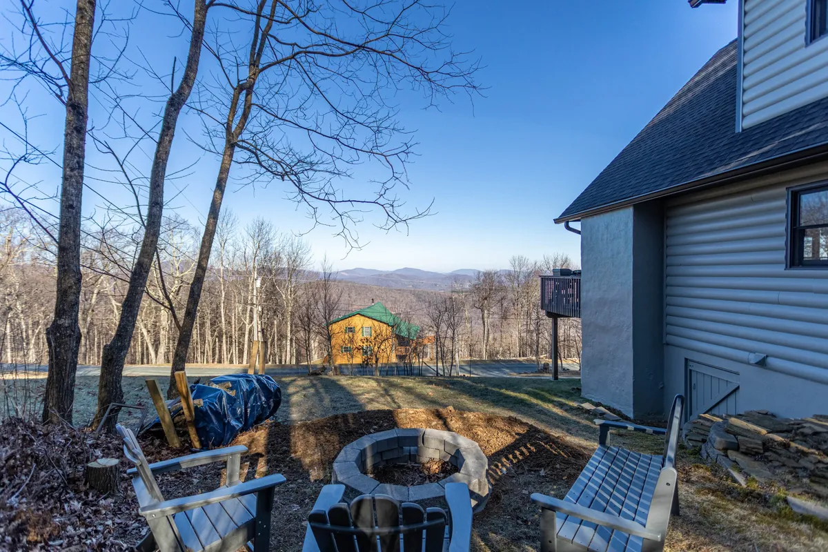 A serene fire pit area with Adirondack chairs arranged around the fire, set against a stunning mountain backdrop. The scene exudes coziness and invites guests to gather for stories, s’mores, or stargazing in the fresh mountain air. The nearby cabin and open natural surroundings complete the inviting outdoor experience.