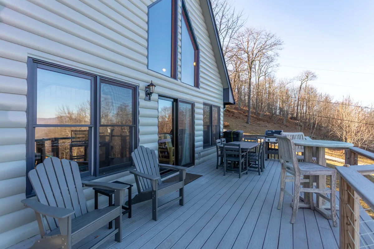 This image shows a spacious outdoor deck furnished with comfortable chairs, large windows offering an indoor-outdoor connection, and an inviting area for outdoor dining. The deck is surrounded by tranquil natural views, creating an inviting space for relaxation or socializing in the fresh mountain air.