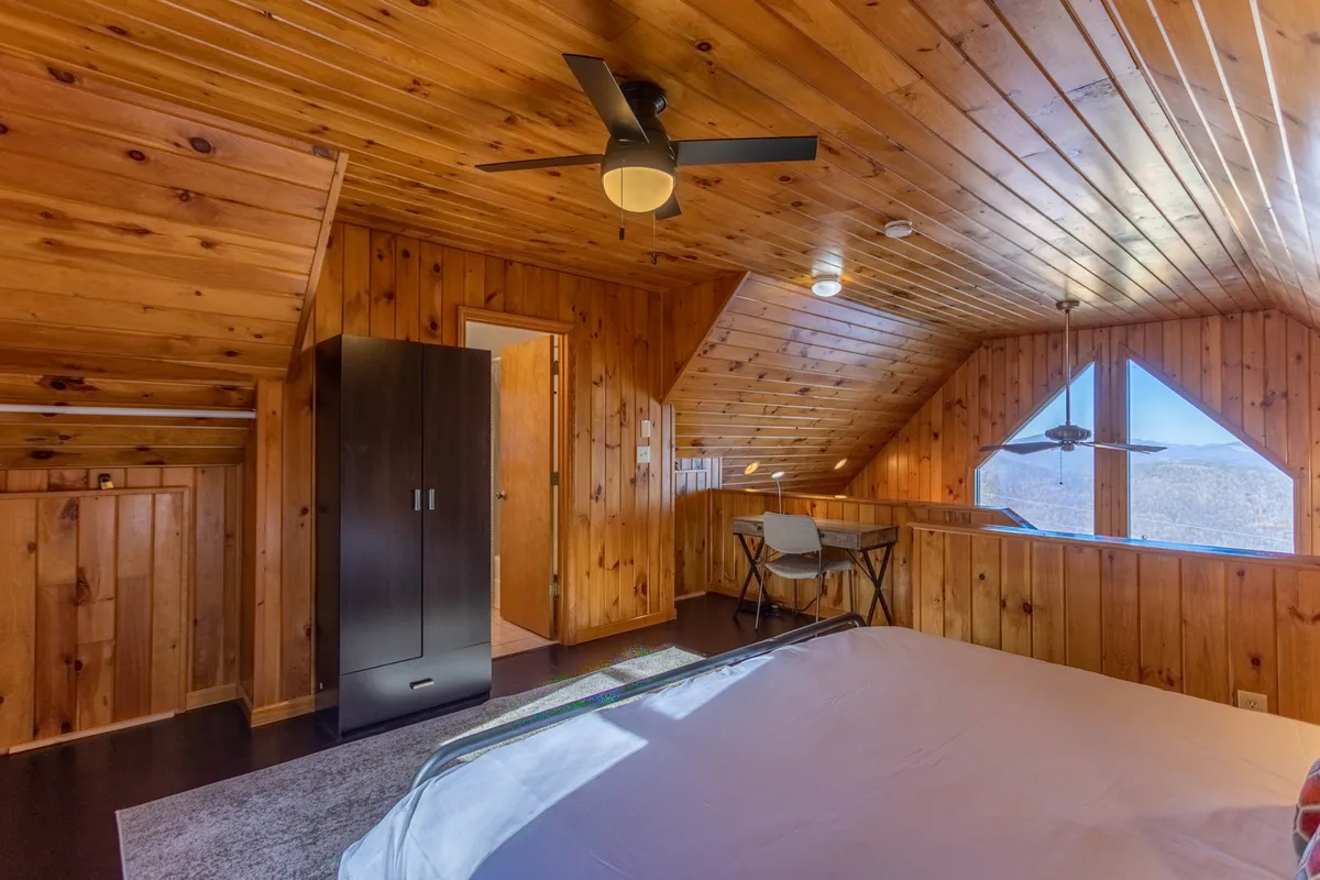 An airy lofted bedroom with a king bed, angled wooden ceilings, large windows letting in natural light, and a modest desk and chair. The elevated position offers privacy and scenic views, evoking a peaceful escape above the main living space.