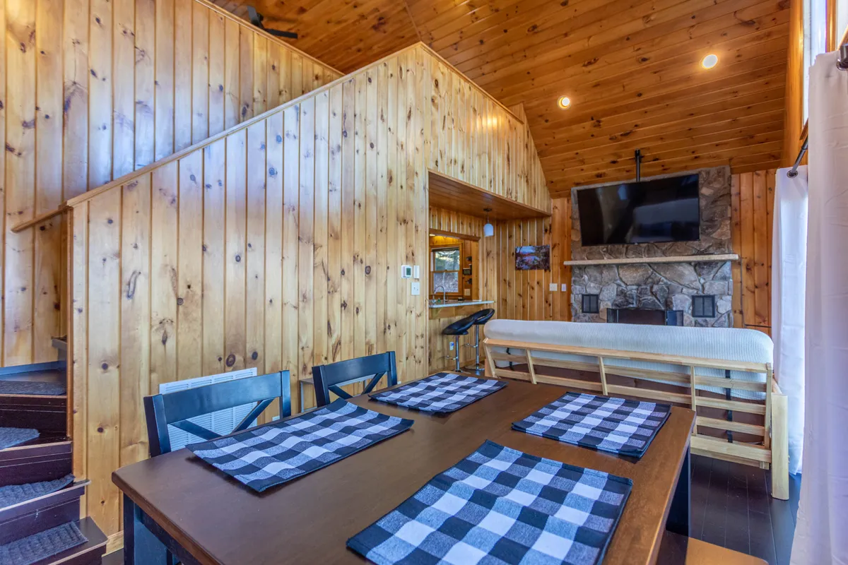 A spacious, open living area features a wooden staircase, dining table set for group meals, a stone fireplace, and a television. The inviting ambiance is perfect for family gatherings, game nights, or relaxing by the fire after a day of exploring nearby Boone.