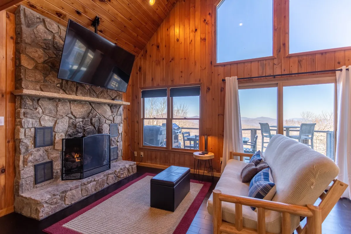 A cozy living room featuring a striking stone fireplace at the center, large windows framing majestic mountain views, a comfortable sofa, and a large TV. The space is warmly lit and exudes an inviting, relaxing ambiance perfect for gatherings or quiet nights in.