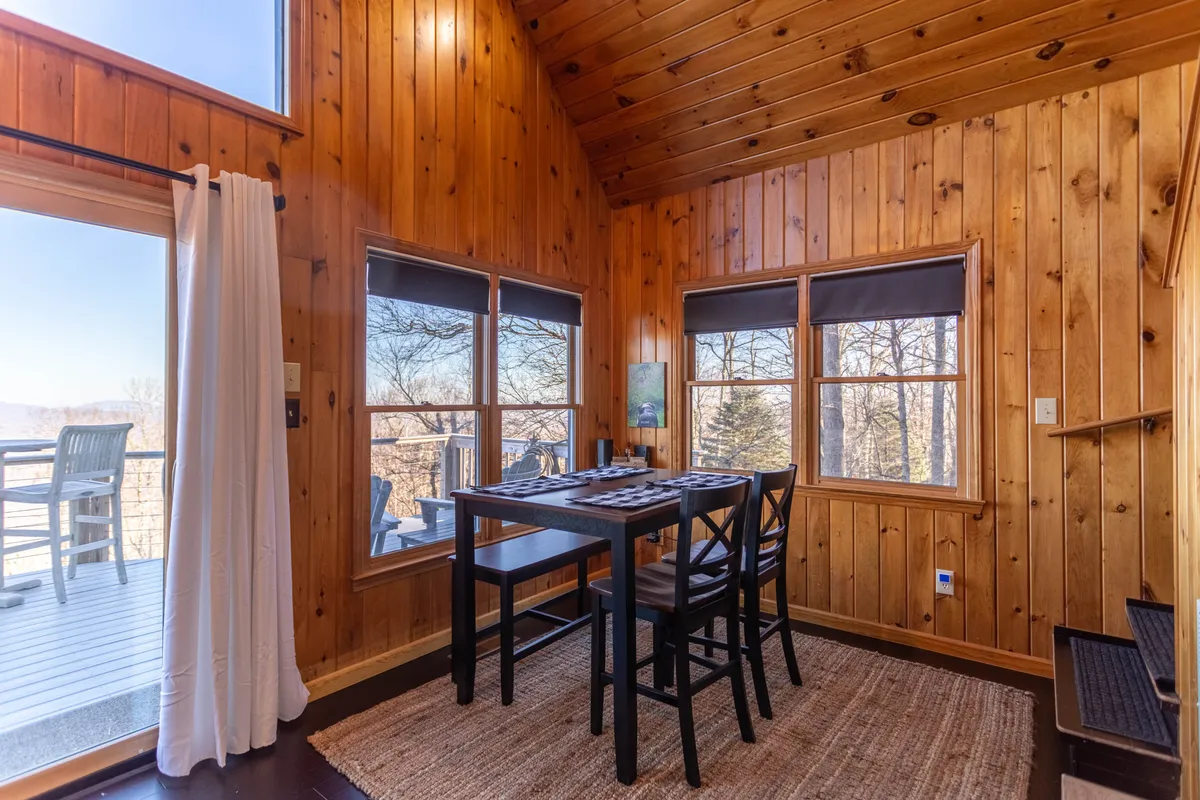 A cozy dining area with a wooden table, black chairs, woven rug, and, most importantly, expansive windows framing a view of the outdoors. The natural light pouring in highlights the comfortable work and gathering space, with the mountains subtly visible through the glass.