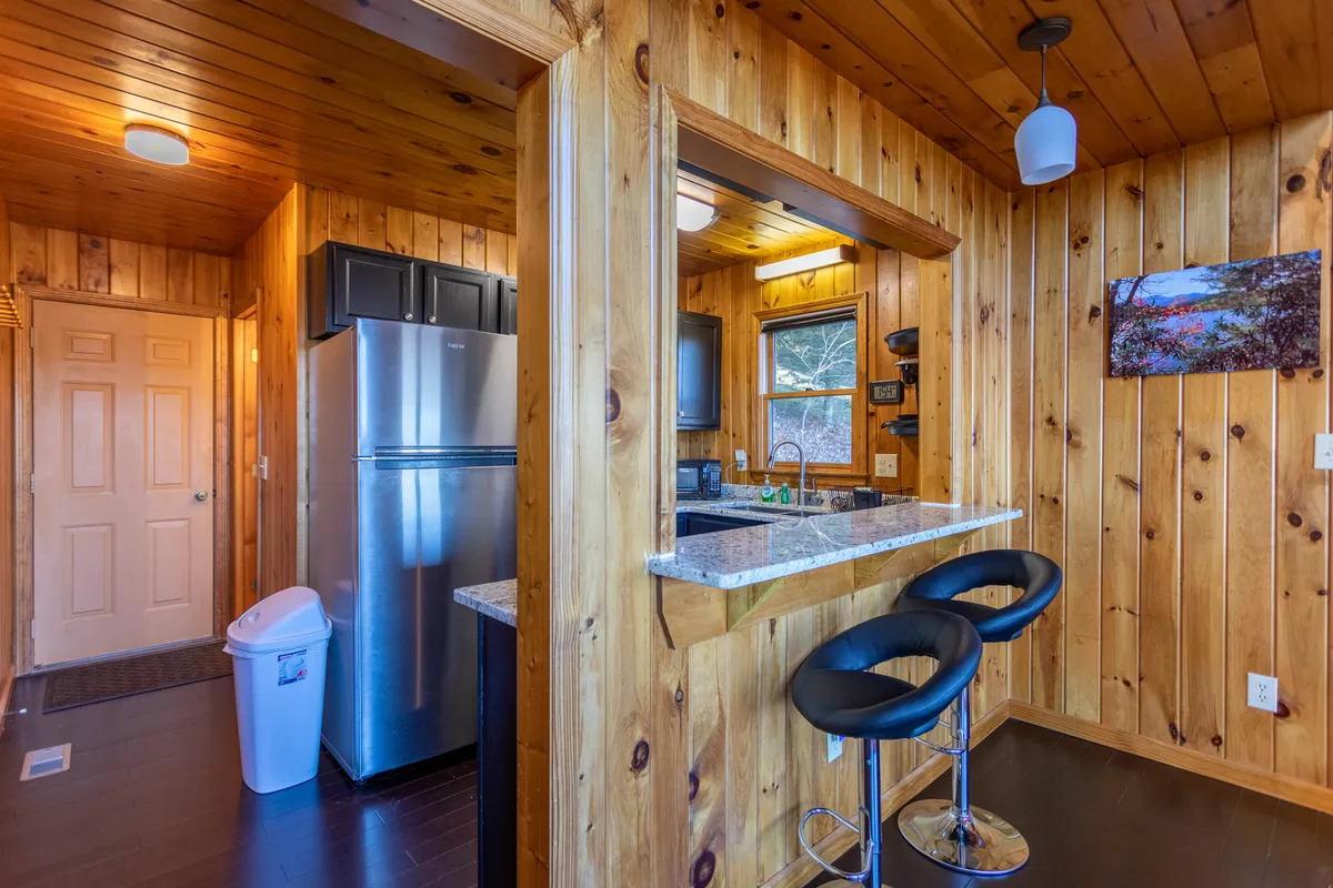 A close-up view of a modern kitchen with gleaming granite countertops, sleek appliances, bar stools, wooden walls, and a window revealing a glimpse of nature outside. The scene suggests a space ideal for cooking, socializing, and enjoying intimate meals.
