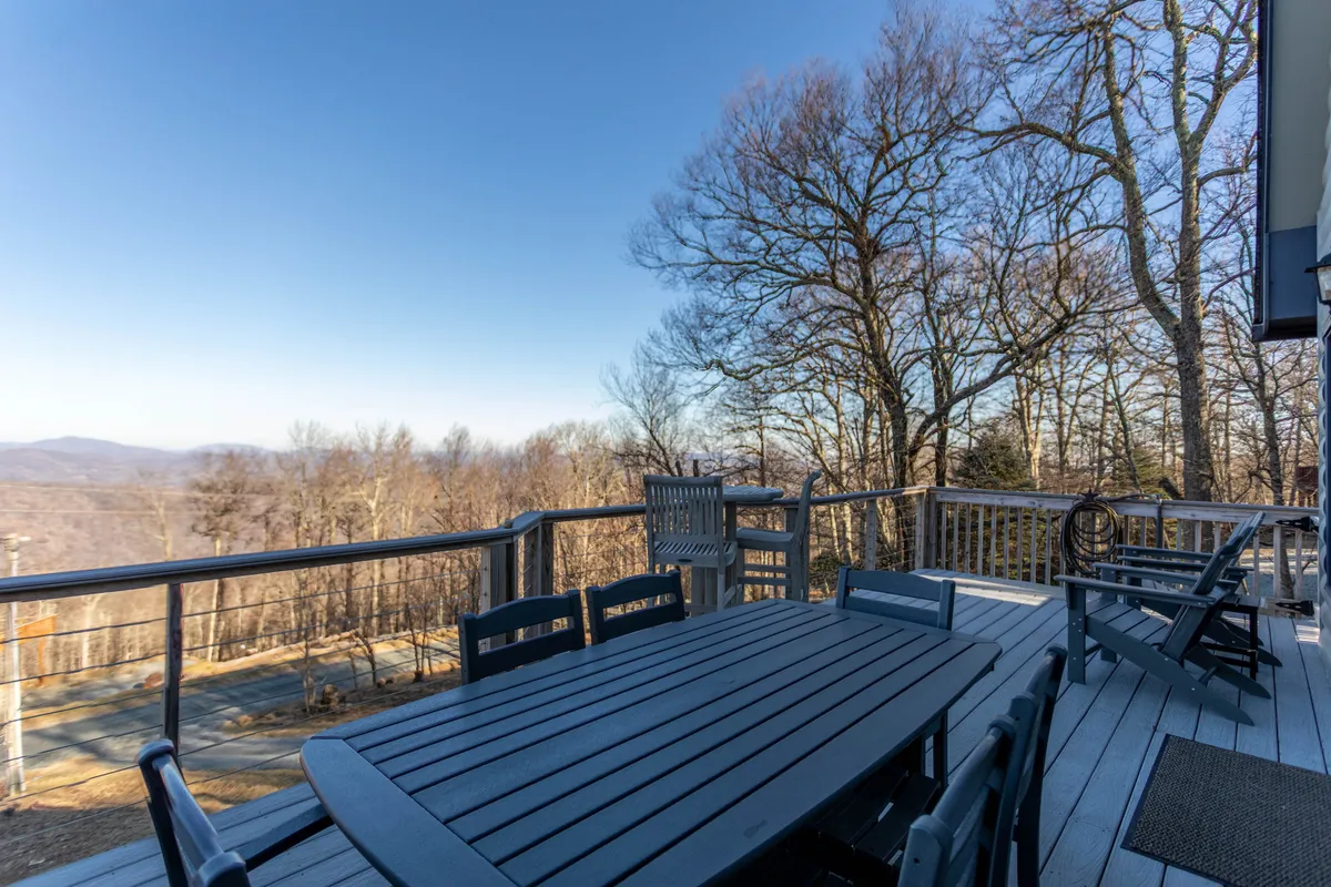 An inviting outdoor dining table and wooden chairs sit on a spacious deck, overlooking breathtaking mountain vistas. The deck is designed for relaxation and social gatherings, with a sturdy railing framing the extraordinary view across the High Country.