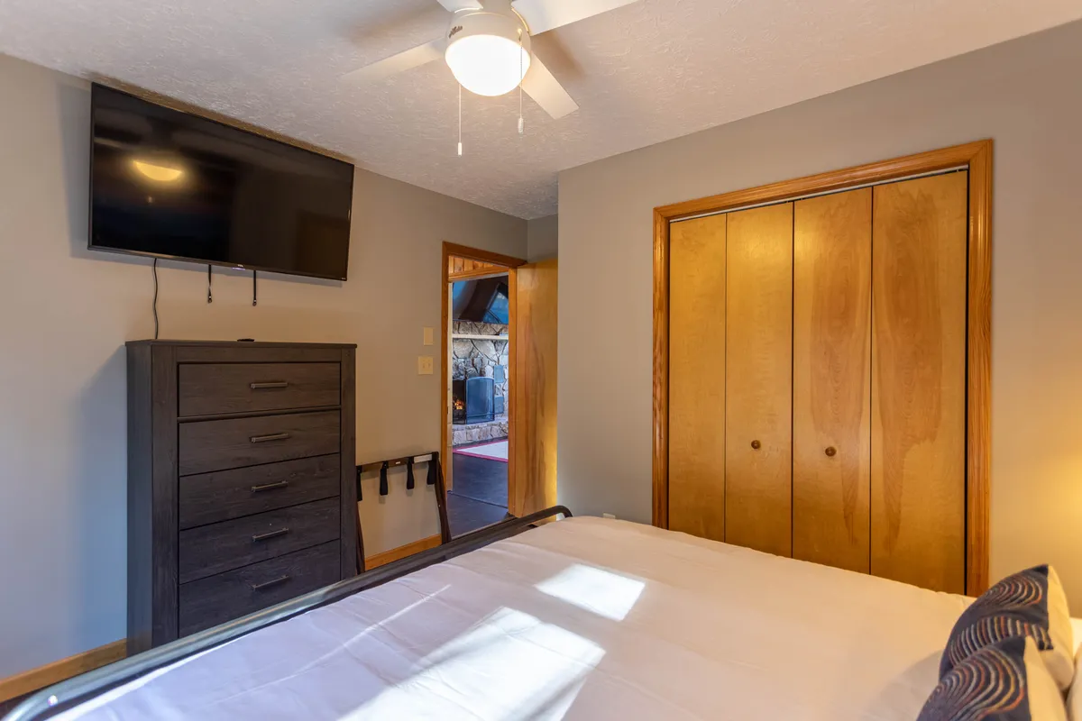 A cozy bedroom featuring a king-size bed, dresser, wall-mounted TV, and closet doors. Soft sunlight filters through a window, illuminating the restful, homey atmosphere. The TV is positioned perfectly for relaxing in bed after a long day.