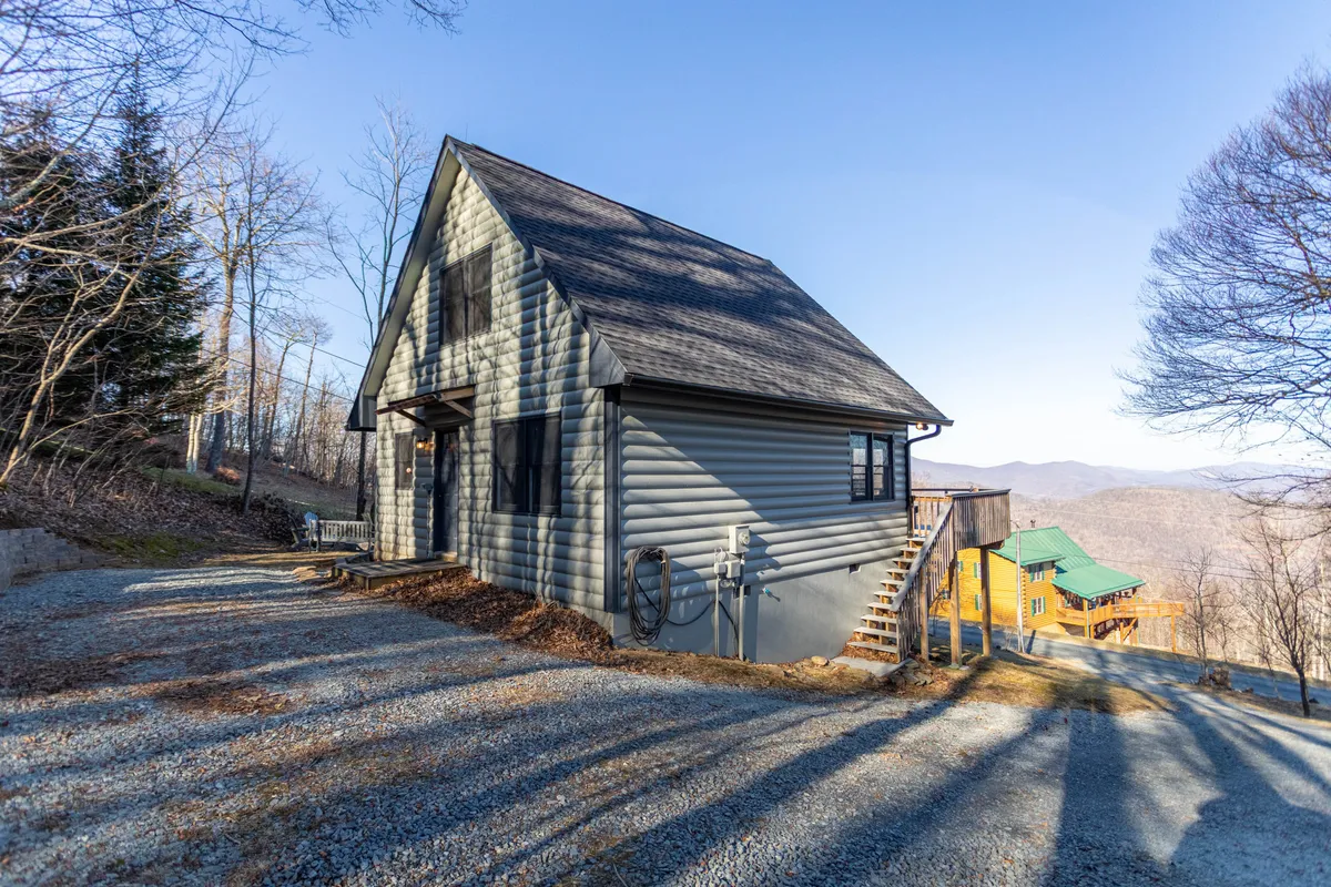 This exterior image displays the house situated against a backdrop of mountains, with the gravel driveway and the front entryway clearly visible. The peaceful setting immediately conveys arrival in a private mountain retreat, and the driveway hints at the need for AWD/4WD access.