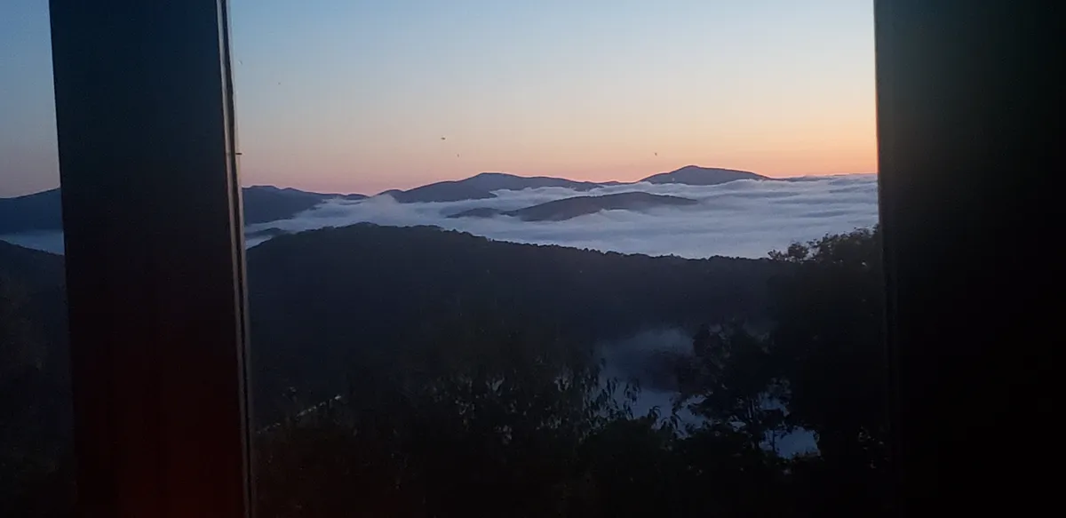 A breathtaking, wide-angle view of misty mountains at sunset as seen from the property. The photo captures rolling peaks shrouded in fog with the warm colors of the setting sun painting the sky. No visible structures, just pure mountain landscape—inviting viewers to imagine themselves immersed in this tranquil natural setting.